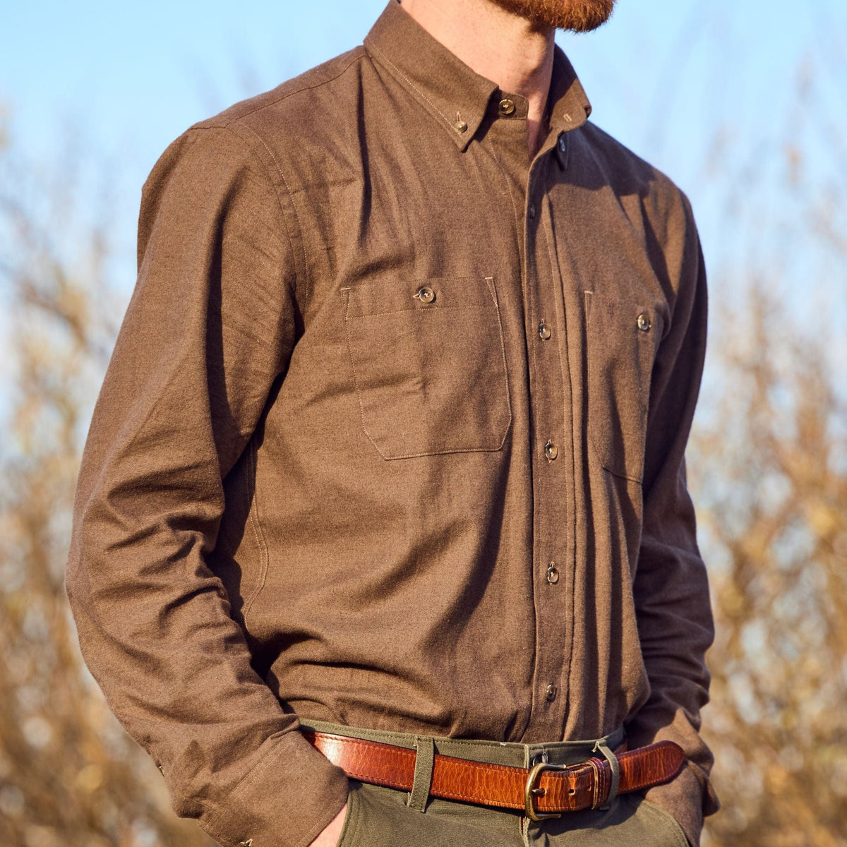Someone stands outdoors in sunlight wearing a Tom Beckbe Dixon Brushed Cotton Twill Shirt, brown belt, and green pants.