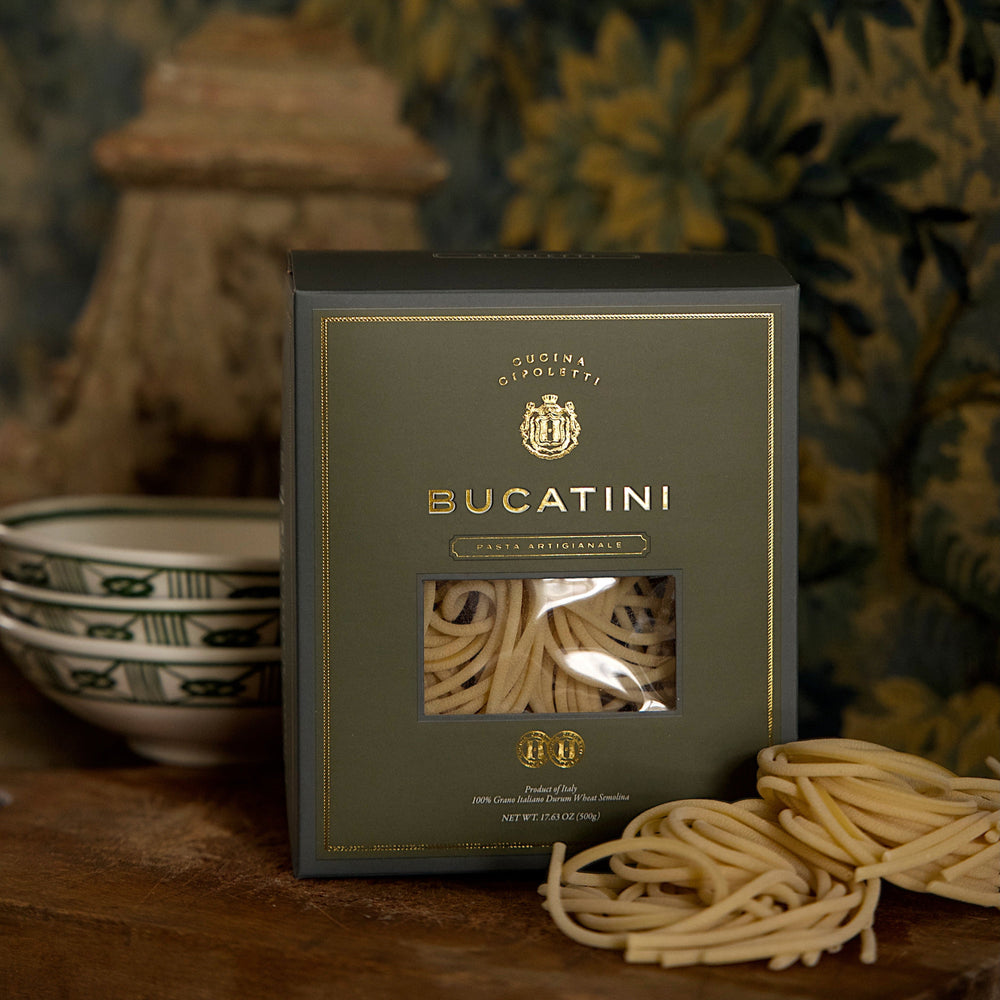 Front view of Bucatini boxed pasta showcasing gold-embossed dark green packaging with window revealing coiled pasta and loose nests on a wooden table