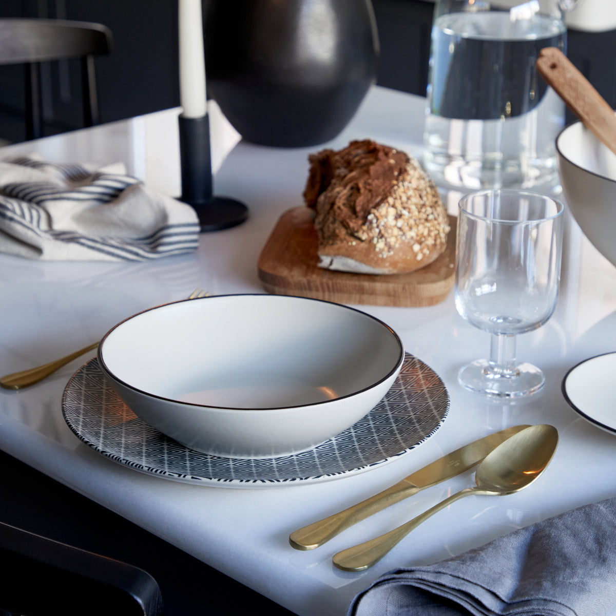 Gold flatware arranged beside a place setting with patterned plates, glassware, bread, and folded napkins