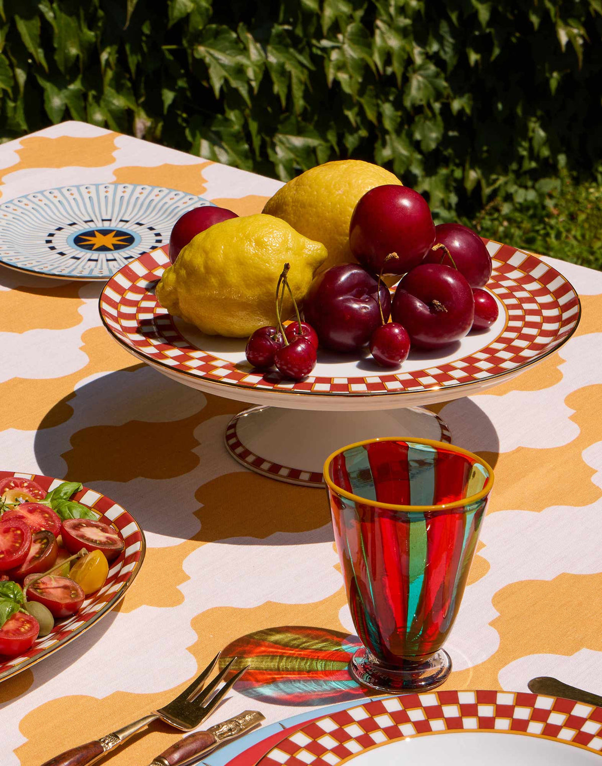 Angled view of Cake Stand in Apollo Mattone with Sun God inspired print 18K gold rim and fruit on a patterned table
