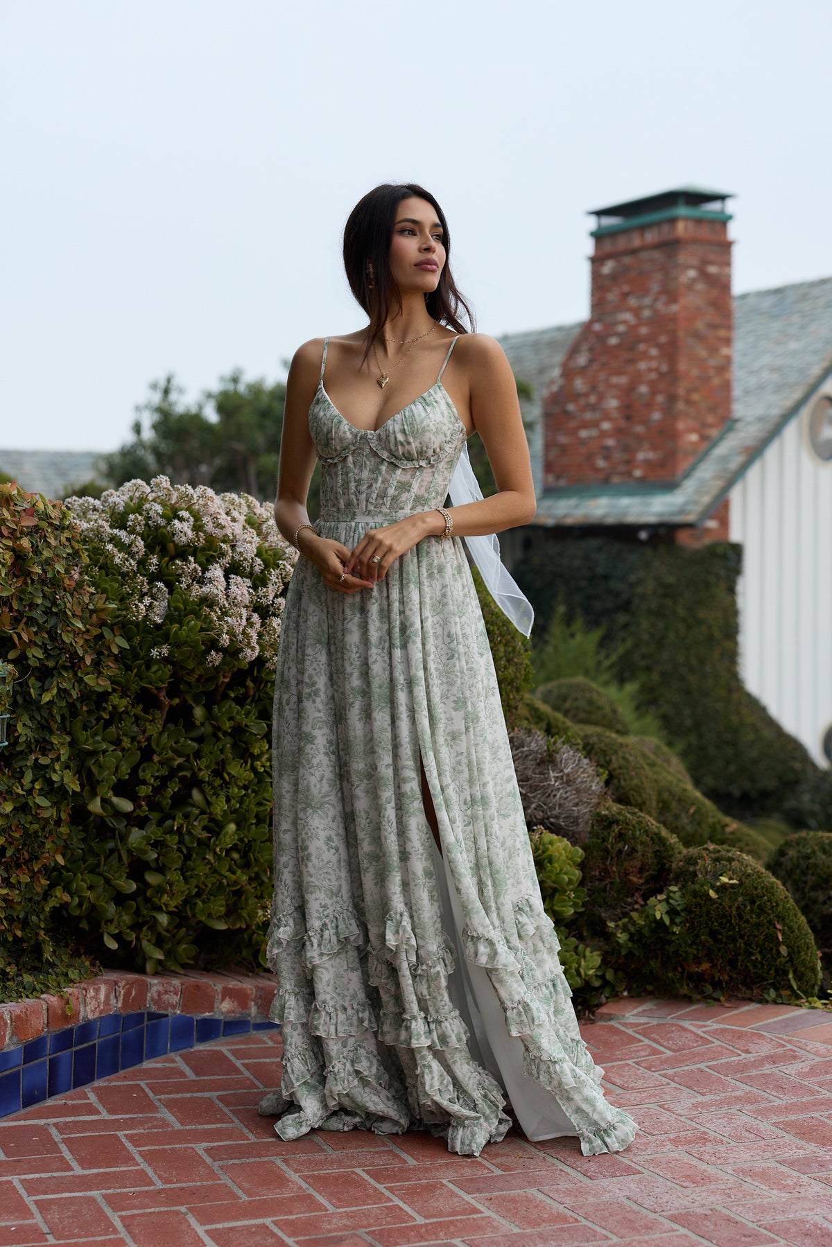 Front view of Carmen Dress in sage toile with corset bodice, side slit, and layered ruffle hem in a garden courtyard