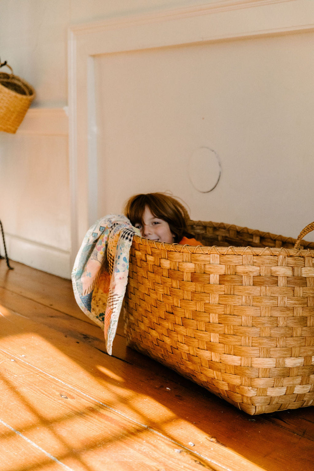 Child peeking from inside a woven Gloucester basket with a patterned quilt draped over the rim on a sunlit wood floor