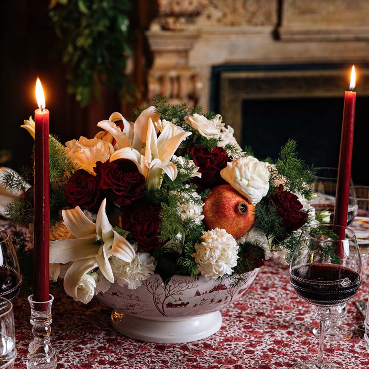 Front view of Country Estate Winter Frolic 13” centerpiece bowl showcasing footed red-and-white scenic pattern, filled with white lilies, red roses and a pomegranate on a holiday table