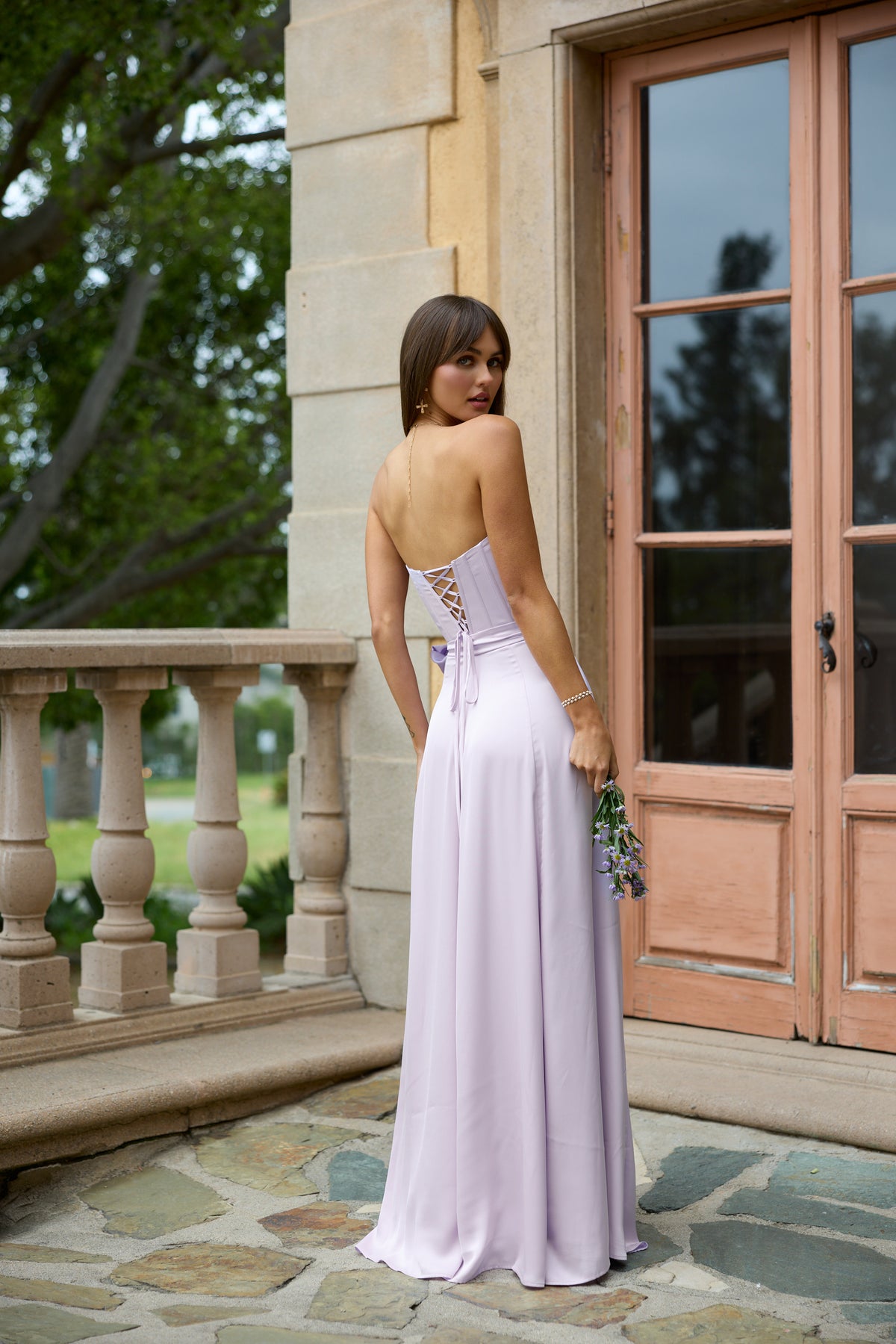 Back view of Cecilia Dress in Lavender Fog showing lace-up corset back and flowing floor-length skirt on a stone terrace