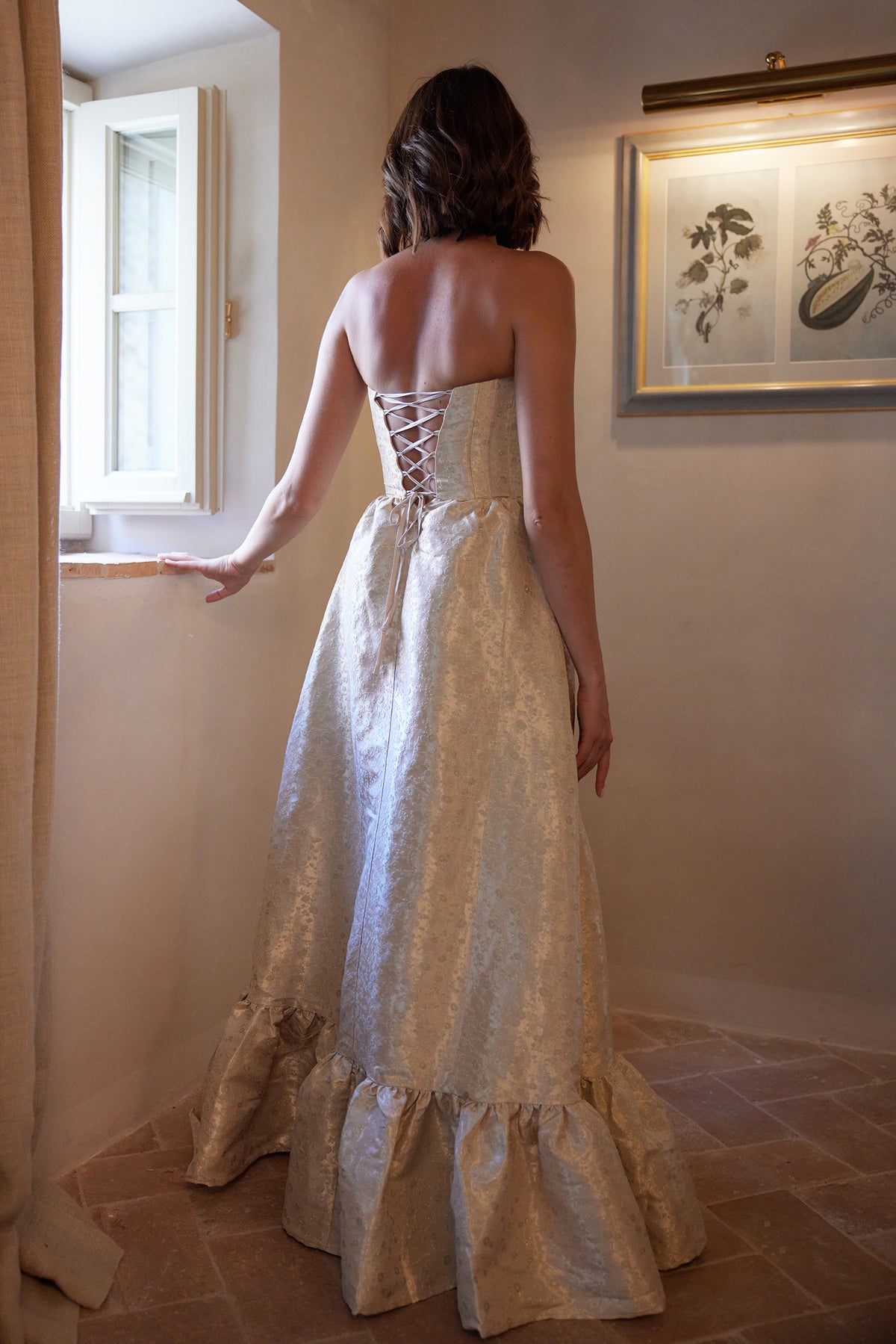 Back view of The Charlotte Dress in Taupe Duchess showcasing its laced corset back and gathered hem in an indoor setting