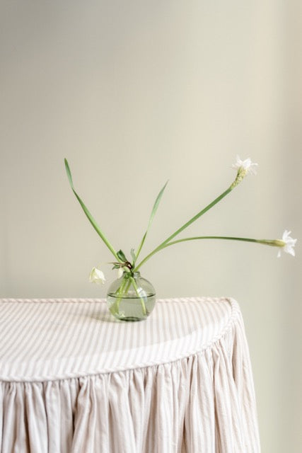 Front view of clear glass bud vase holding three long green stems with white blooms on a pleated striped tablecloth