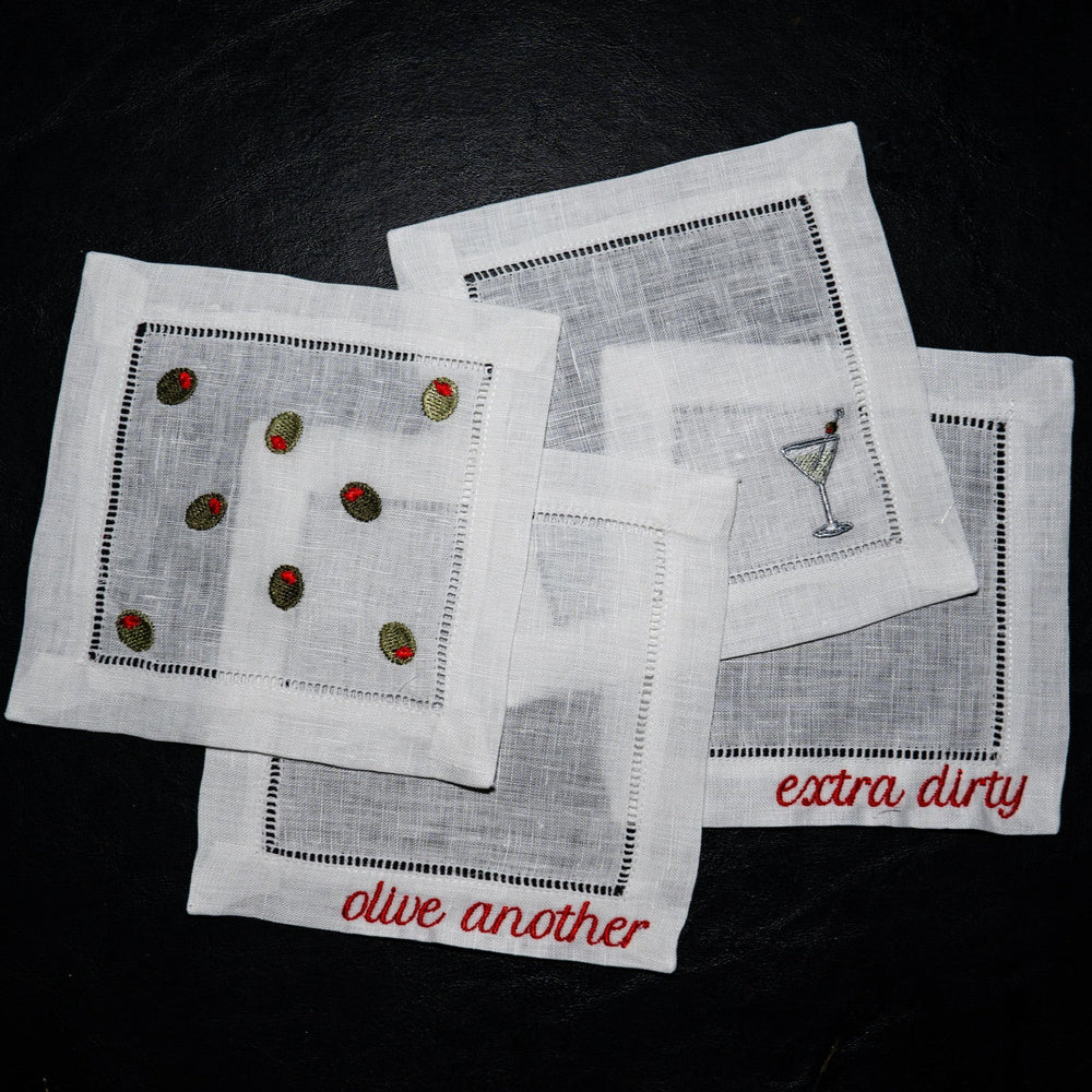 Flat lay of linen martini cocktail napkins set of 4 embroidered with olives, a martini glass, and sayings olive another and extra dirty on black background