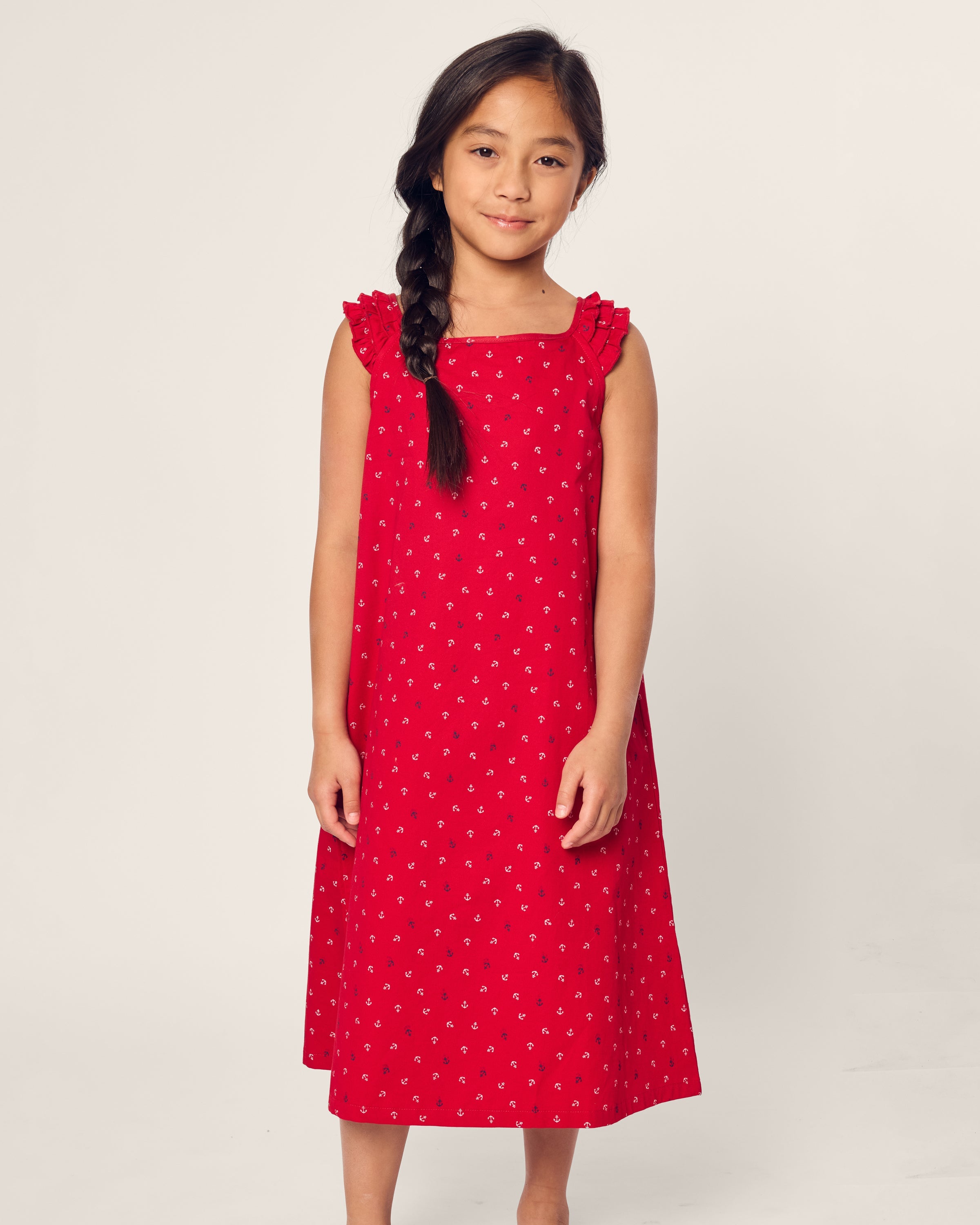 A girl with long dark hair in a braid wears the Petite Plume Girls Twill Amelie Nightgown in Anchors Away. She stands barefoot against a light background, smiling slightly with her hands at her sides.
