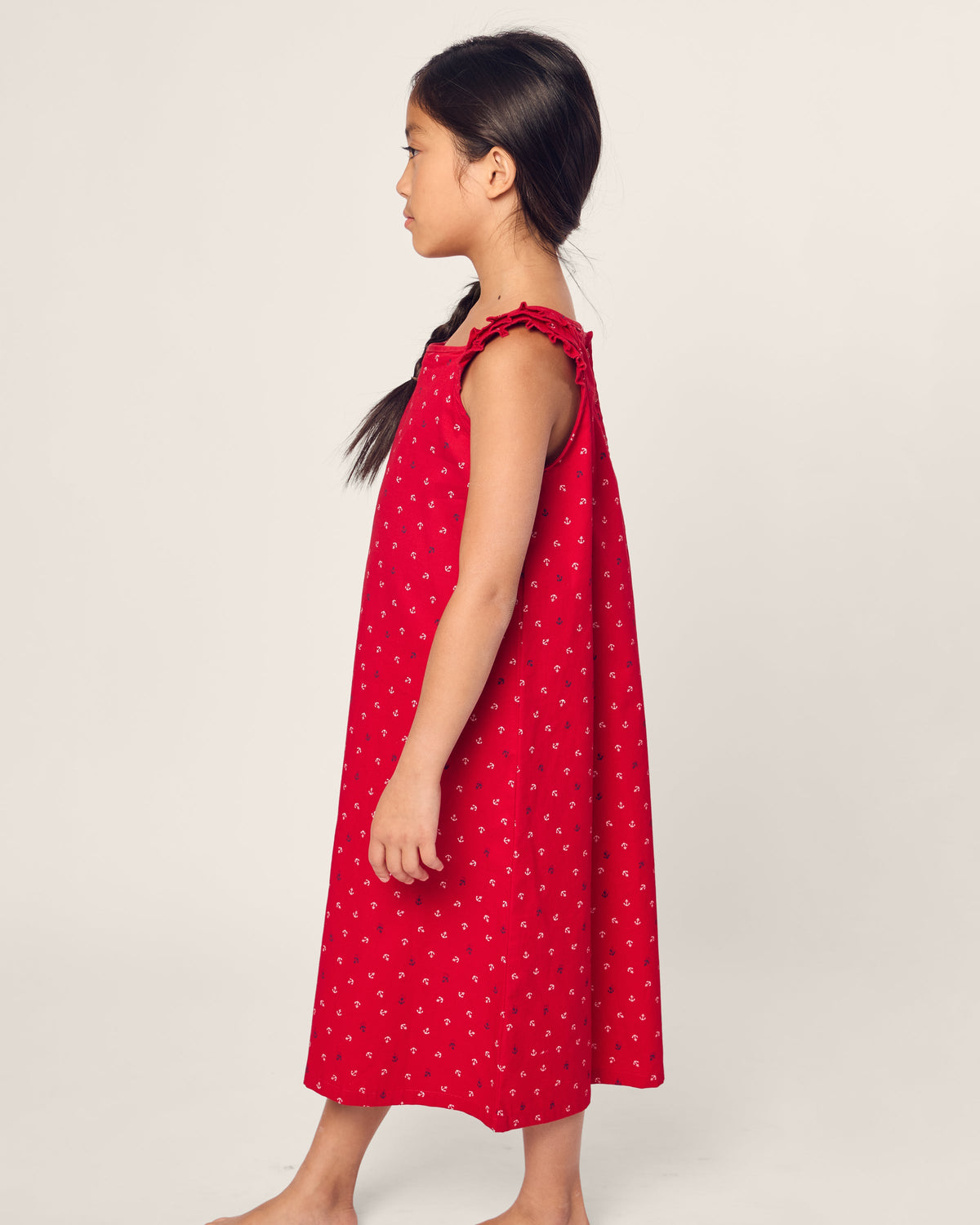 A young girl stands in profile against a plain background, wearing the Girls Twill Amelie Nightgown in Anchors Away by Petite Plume. The luxurious sleepwear features ruffled straps, a nautical print with small white patterns on red fabric, and her hair is styled in a long braid. She is barefoot.