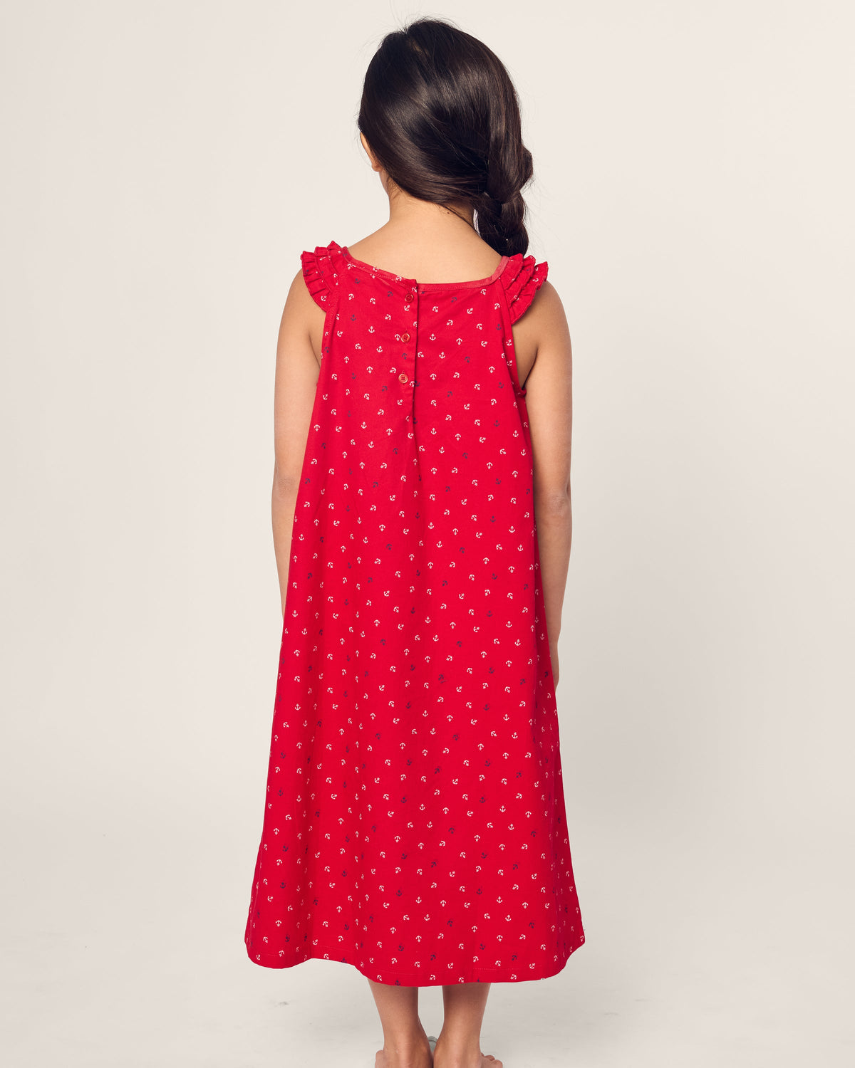 A young girl stands barefoot with her back to the camera, wearing a Petite Plume Girls Twill Amelie Nightgown in Anchors Away. Her long dark hair is styled in a side braid, set against a plain, light-colored background.