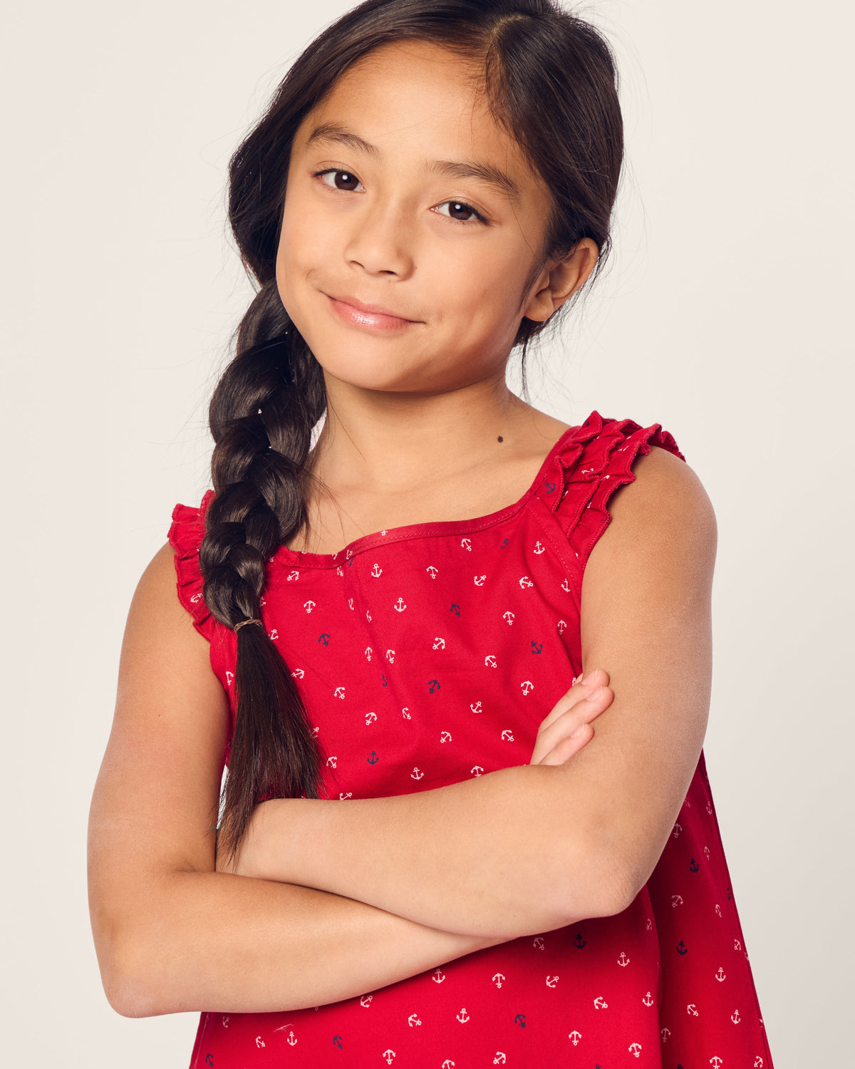 A young girl with long dark hair braided to one side stands confidently with her arms crossed, wearing the Petite Plume Girls Twill Amelie Nightgown in Anchors Away. The lightweight woven cotton enhances her relaxed look as she smiles subtly, gazing at the camera.