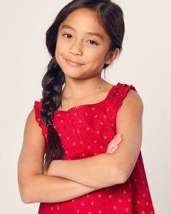 A young girl with long dark hair braided to one side stands confidently with her arms crossed, wearing the Petite Plume Girls Twill Amelie Nightgown in Anchors Away. The lightweight woven cotton enhances her relaxed look as she smiles subtly, gazing at the camera.
