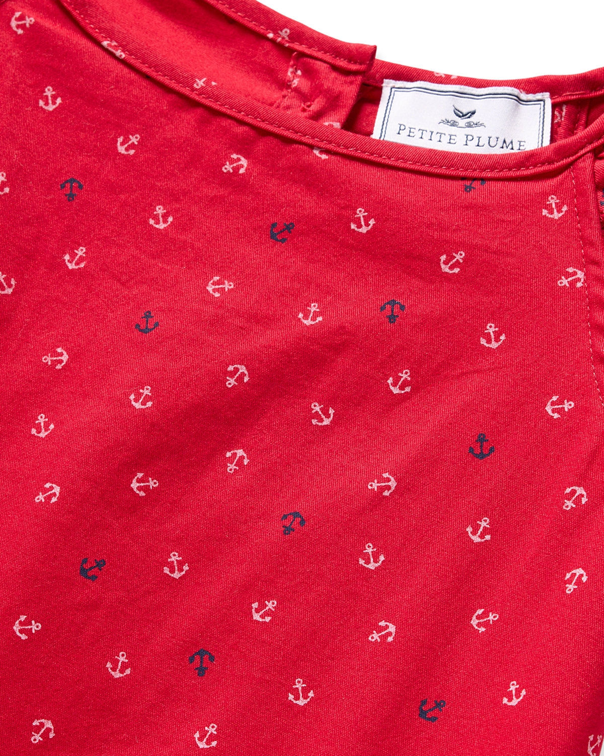 Close-up of a red fabric with small white and navy anchors, radiating nautical charm. The label reads Petite Plume, indicating the luxurious Girls Twill Amelie Nightgown in Anchors Away, crafted from lightweight woven cotton for ultimate comfort.