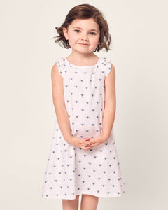 A young girl with short brown hair smiles in a sleeveless Girls Twill Amelie Nightgown in Buzzing Bees by Petite Plume. Her hands are clasped, and she stands against a plain, light background.