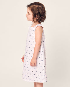 A young child with wavy brown hair stands gracefully in profile against a light background, wearing the Girls Twill Amelie Nightgown in Buzzing Bees by Petite Plume.