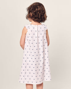 A young child with curly hair stands facing away, wearing Petite Plumes Girl’s Twill Amelie Nightgown in Buzzing Bees, a sleeveless white cotton nightgown with small dark blue bees pattern. The background is neutral light gray.