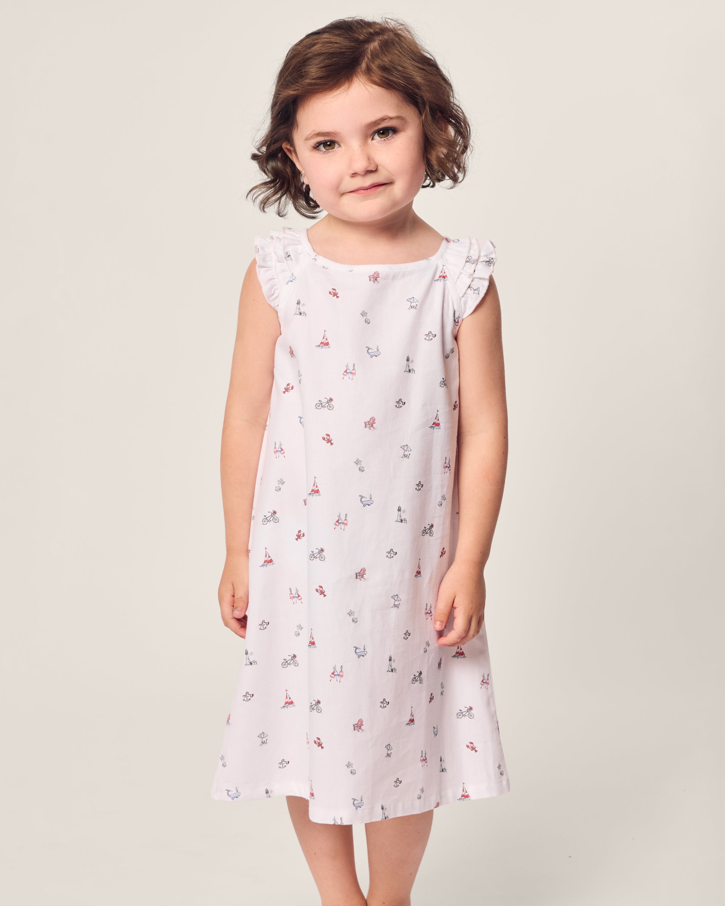 A young child with brown hair wears a Petite Plume Girls Twill Amelie Nightgown in Coastal Charms, made of lightweight cotton. The gown, featuring small red and blue patterns, beautifully contrasts against the plain light background.