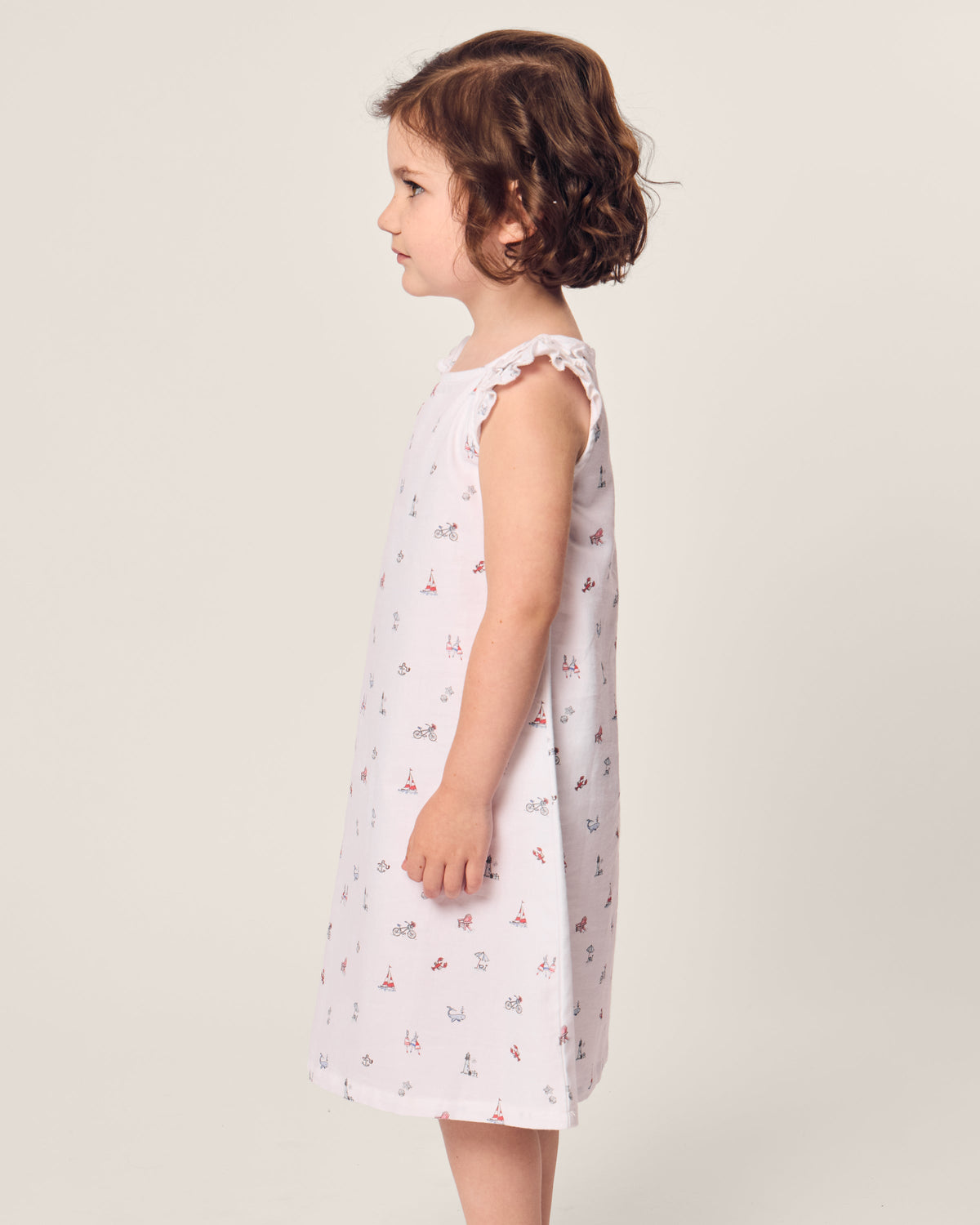 A young girl with short brown hair stands in profile, wearing Petite Plumes Girls Twill Amelie Nightgown in Coastal Charms, featuring small scattered patterns. She is set against a plain, light background.