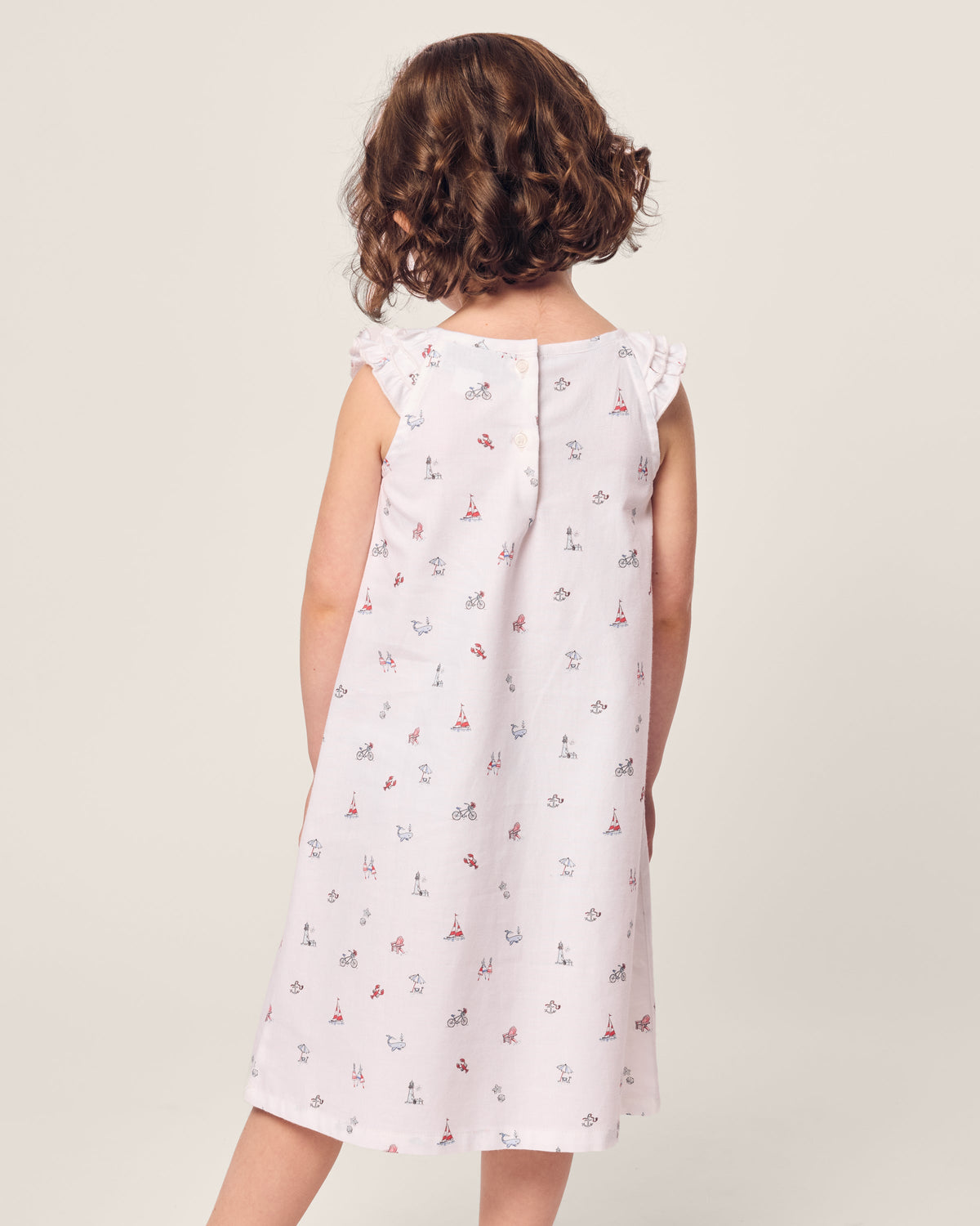 A young girl with curly brown hair stands with her back to the camera, wearing Petite Plumes Girls Twill Amelie Nightgown in Coastal Charms—a sleeveless white dress with red and blue sailboat and flower patterns. Made of lightweight cotton, it contrasts elegantly against the plain light-colored background.