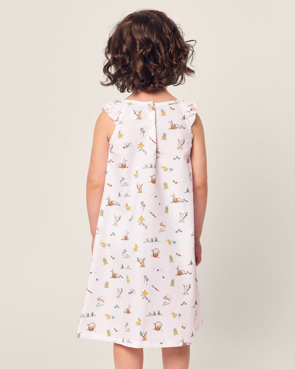 A young girl with curly hair stands facing away, wearing the sleeveless Girls Twill Amelie Nightgown in Easter Frolic by Petite Plume. The soft neutral background highlights her casual and whimsical attire.