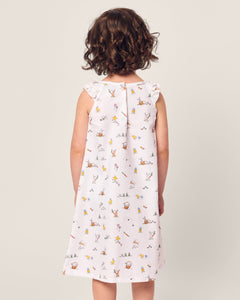 A young girl with curly hair stands facing away, wearing the sleeveless Girls Twill Amelie Nightgown in Easter Frolic by Petite Plume. The soft neutral background highlights her casual and whimsical attire.