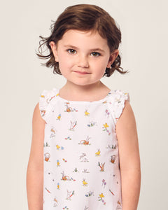 A young girl with short brown hair stands against a light background, wearing a Petite Plume Girls Twill Amelie Nightgown featuring Easter Frolic prints of animals and plants, looking directly at the camera.