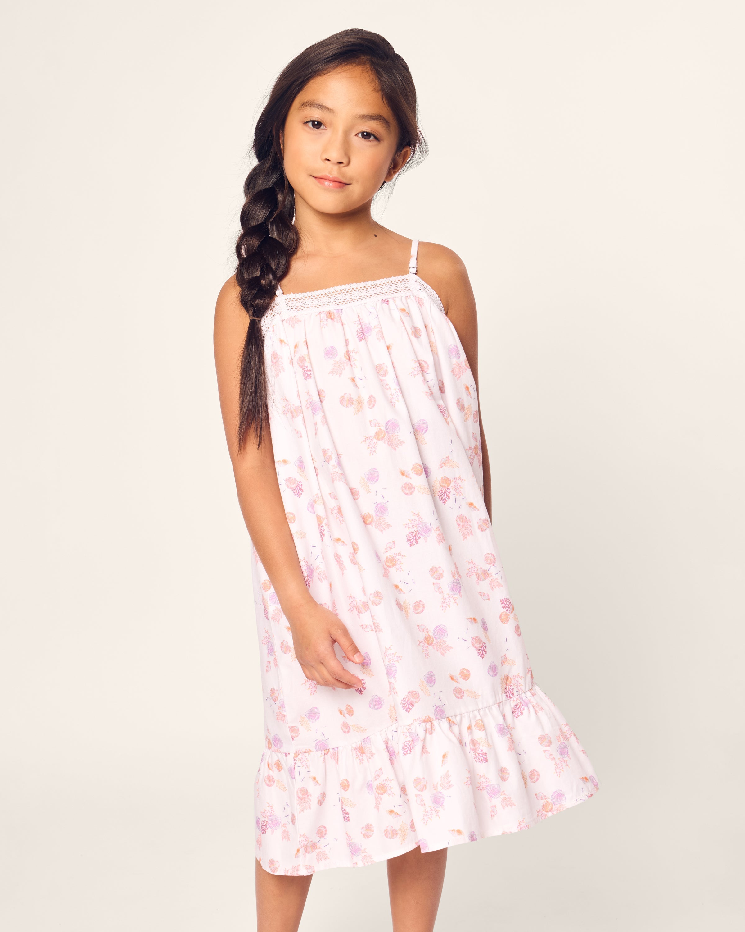 A girl with long braided hair wears Petite Plumes Girls Twill Lily Nightgown in Seashell Splendor. The nightgown, made of white yarn-dyed cotton with pink floral patterns, contrasts against a plain light background as she looks at the camera with a slight smile.