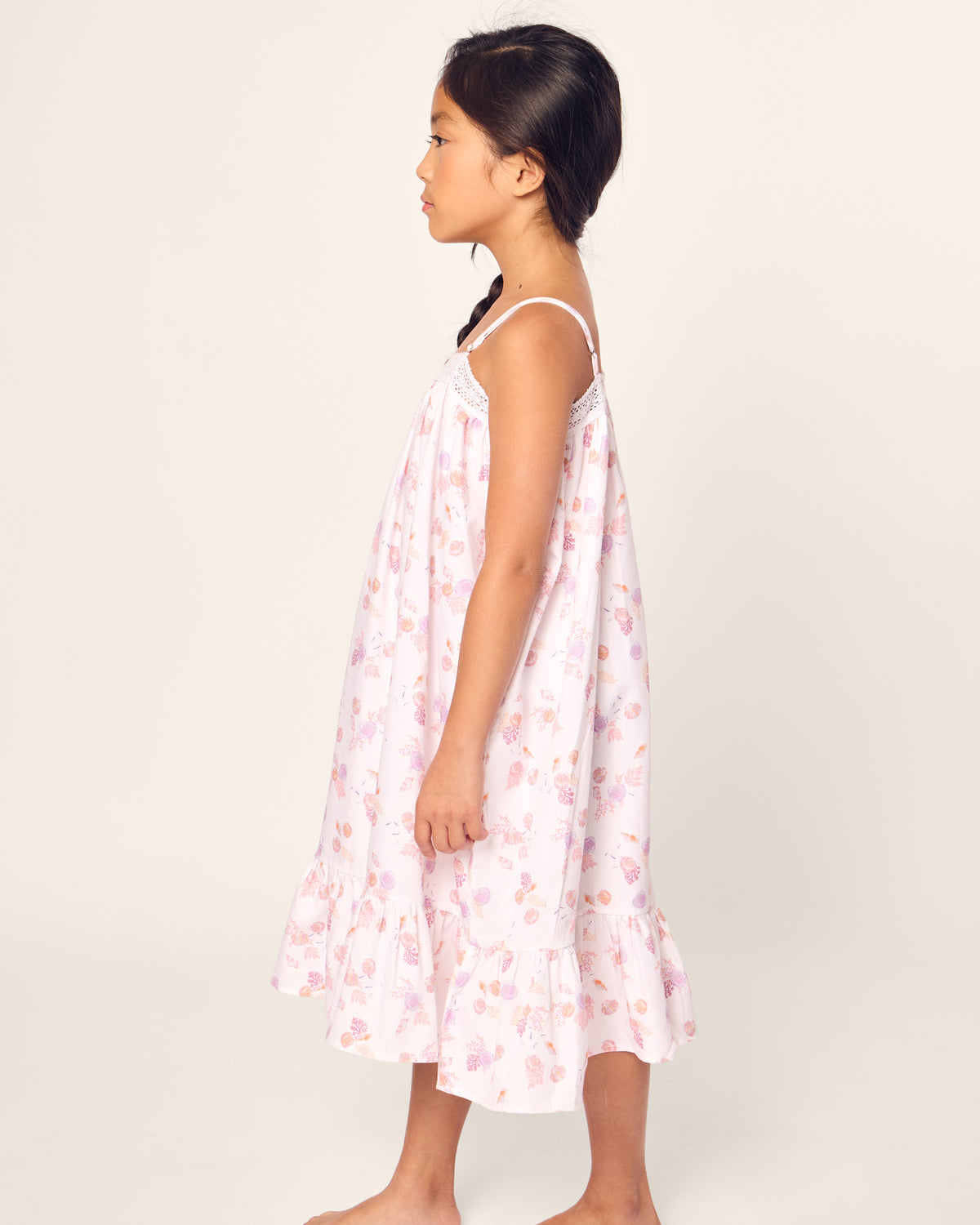 A girl in a sleeveless pink floral dress with a ruffled hem, her dark braid hanging down as she stands sideways, exudes the charm of luxurious sleepwear akin to Petite Plumes Girls Twill Lily Nightgown in Seashell Splendor against a plain background.