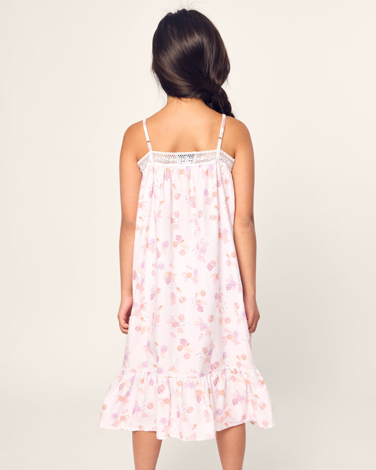 A girl with long hair stands with her back to the camera, wearing Petite Plumes Girls Twill Lily Nightgown in Seashell Splendor. The lace detailing and ruffled hem evoke luxurious elegance against a light-colored backdrop.