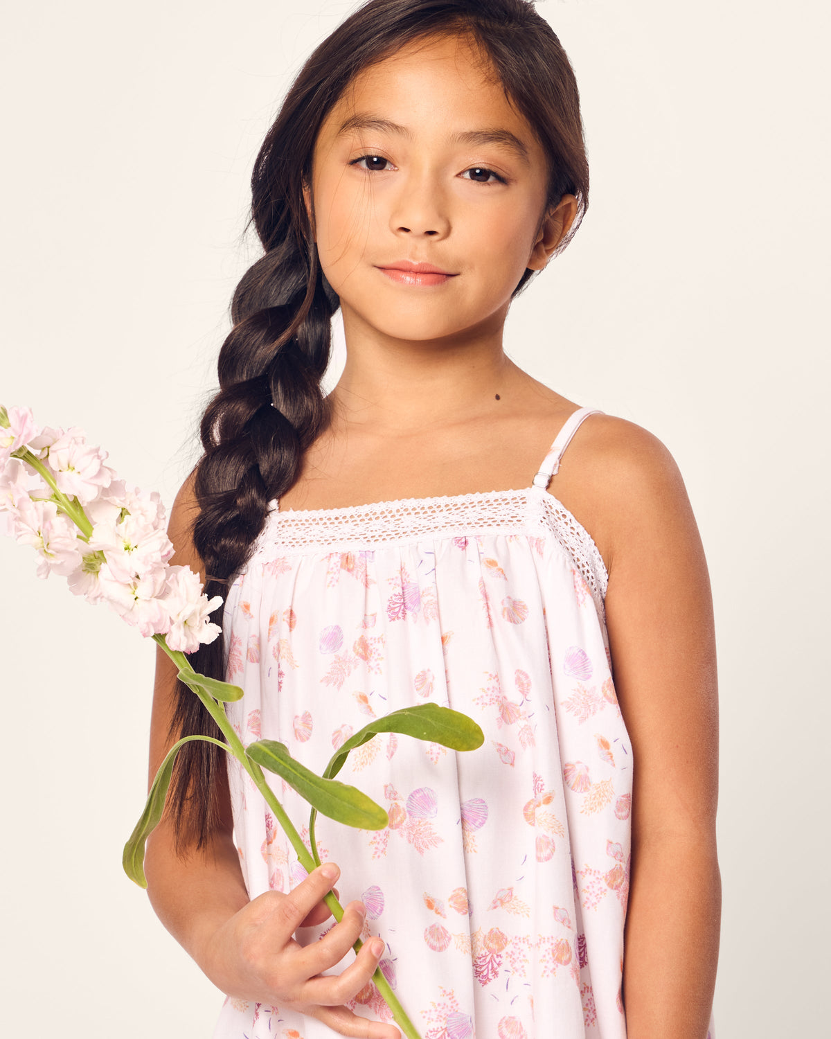 A young girl with long, braided hair holds a pink flower with green leaves, wearing Petite Plumes Girls Twill Lily Nightgown in Seashell Splendor. The background is plain and light-colored.