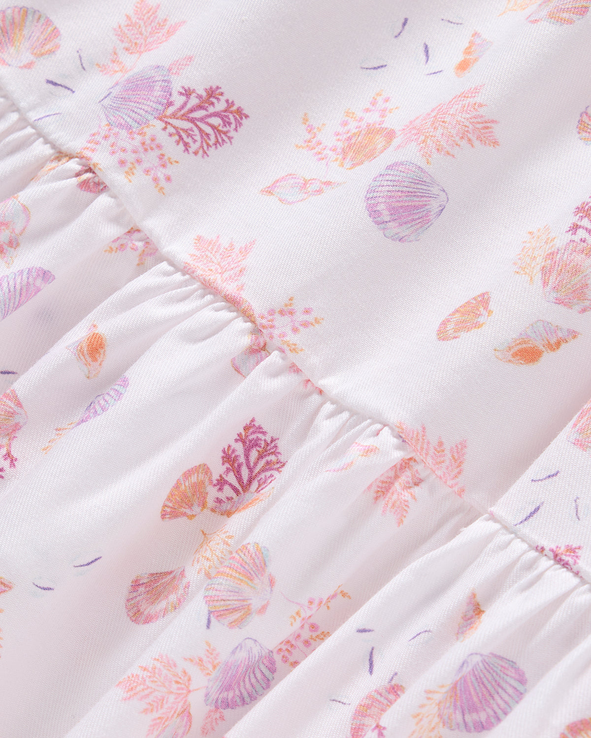 Close-up of the Girls Twill Lily Nightgown in Seashell Splendor by Petite Plume, featuring a light pink backdrop with seashells and coral in pink, orange, and purple. The yarn-dyed cotton is gathered for soft folds and texture, capturing luxurious sleepwear.