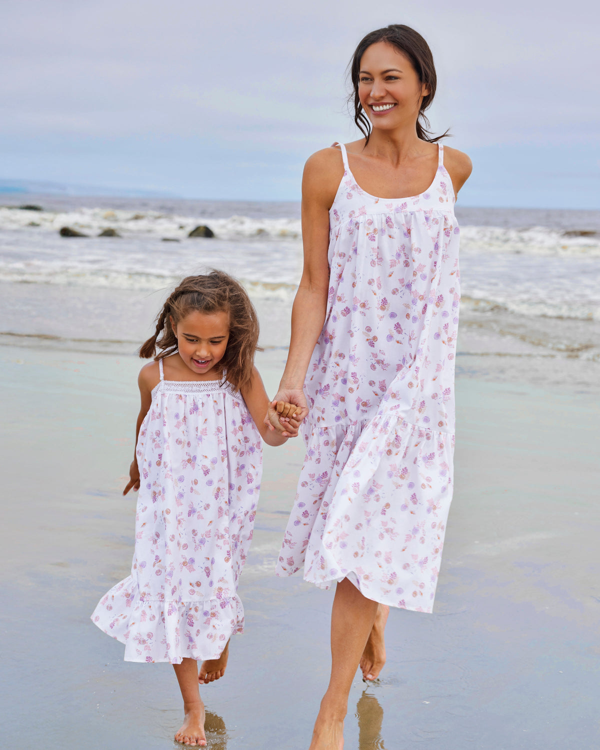 A woman and young girl joyfully stroll barefoot along the beach, holding hands in matching white sundresses adorned with purple floral patterns, echoing the Girls Twill Lily Nightgown in Seashell Splendor by Petite Plume as the waves softly crash behind them.