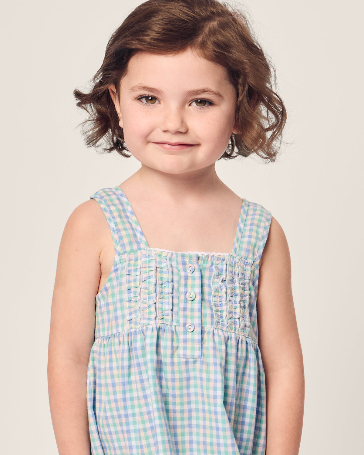 A young girl with short brown hair, wearing the Girls Twill Charlotte Nightgown in Spring Gingham by Petite Plume, smiles gently against a plain, light background.