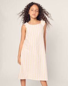 A young girl with curly hair confidently poses in the Petite Plume Girls Twill Amelie Nightgown in Sunny Stripe, a sleeveless cotton dress with yellow vertical stripes and ruffled shoulders against a plain light background.