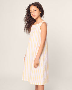 A young girl with curly hair stands in Petite Plumes Girls Twill Amelie Nightgown in Sunny Stripe, a sleeveless, knee-length luxury sleepwear piece made of lightweight cotton. She gazes forward with a neutral expression, exuding effortless elegance against a plain background.