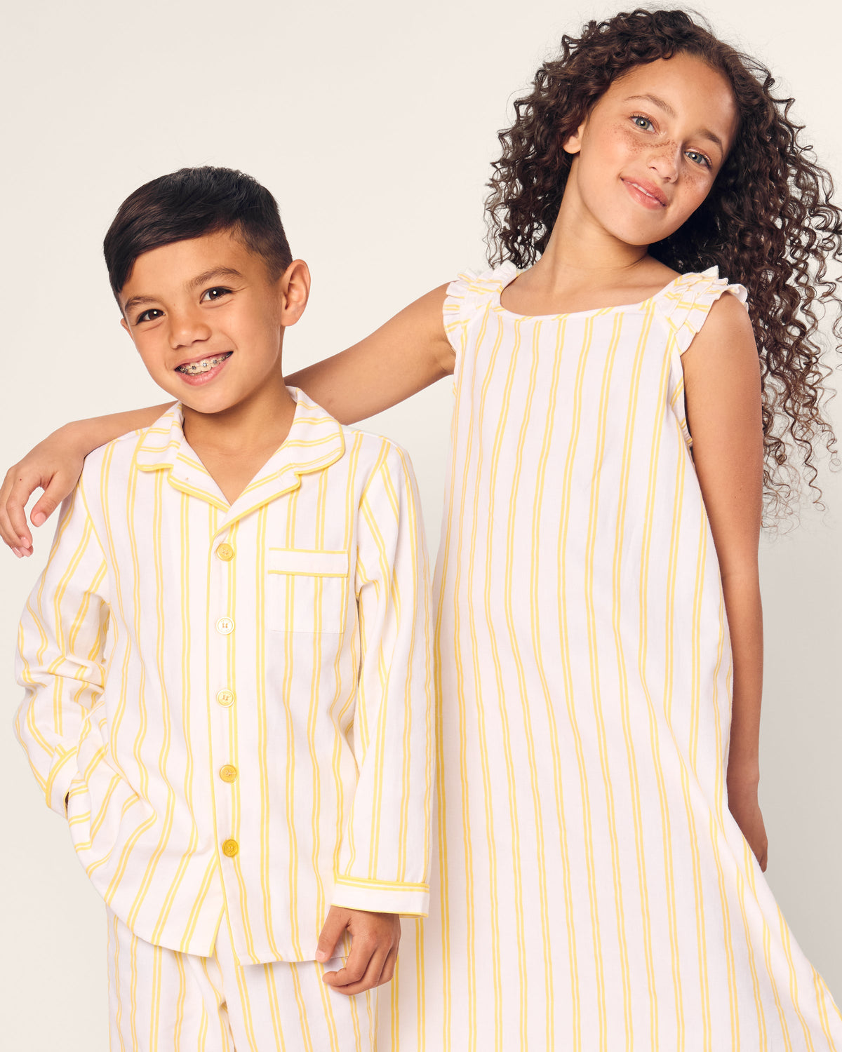 Two children smile in matching luxury sleepwear with yellow and white stripes. The boys PJs feature a collar and buttons, while the girl wears Petite Plumes Girl’s Twill Amelie Nightgown in Sunny Stripe and wraps an arm around his shoulders, set against a plain backdrop.