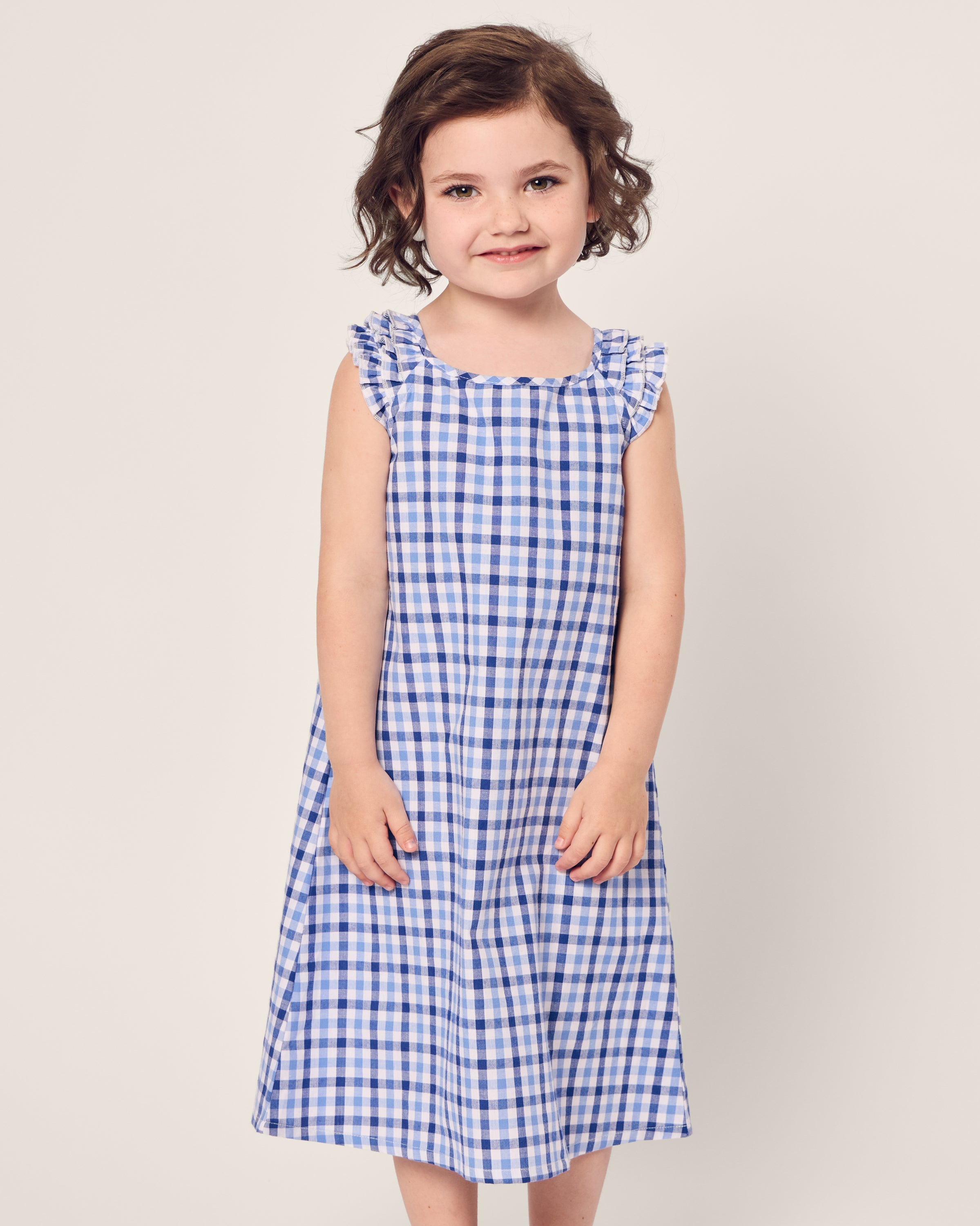 A young girl with short brown hair smiles, wearing the Petite Plume Girls Twill Amelie Nightgown in Royal Blue Gingham. The background is plain off-white.