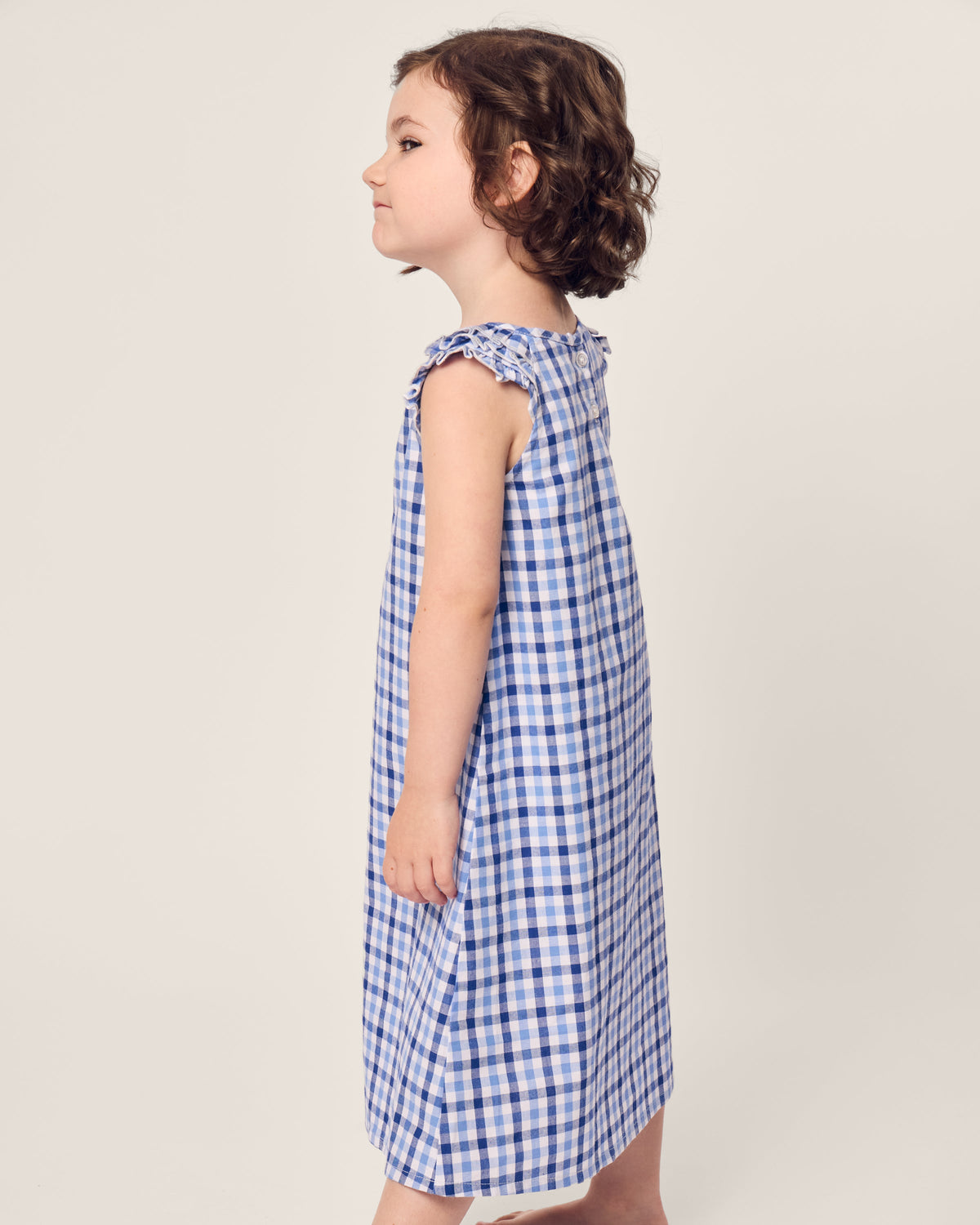 A child with curly brown hair and a calm expression stands sideways against a plain background, wearing the Girls Twill Amelie Nightgown in Royal Blue Gingham by Petite Plume.