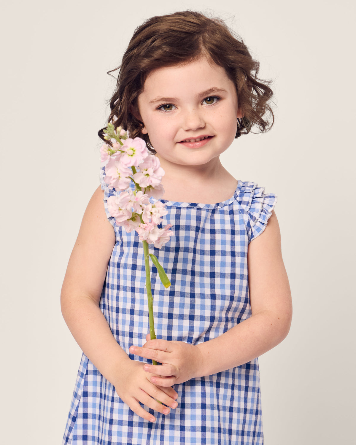 A young child with wavy brown hair stands holding a stem of light pink flowers, wearing Petite Plumes Girls Twill Amelie Nightgown in Royal Blue Gingham, and smiling softly against a plain, light background.