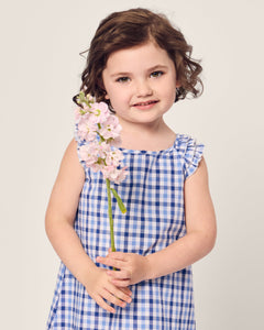 A young child with wavy brown hair stands holding a stem of light pink flowers, wearing Petite Plumes Girls Twill Amelie Nightgown in Royal Blue Gingham, and smiling softly against a plain, light background.