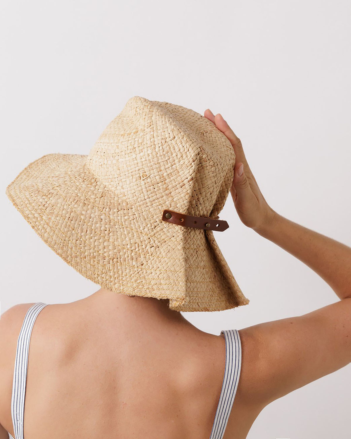 Close-up view of Commando Hat in Natural showcasing raffia weave and adjustable leather tab with a hand holding it