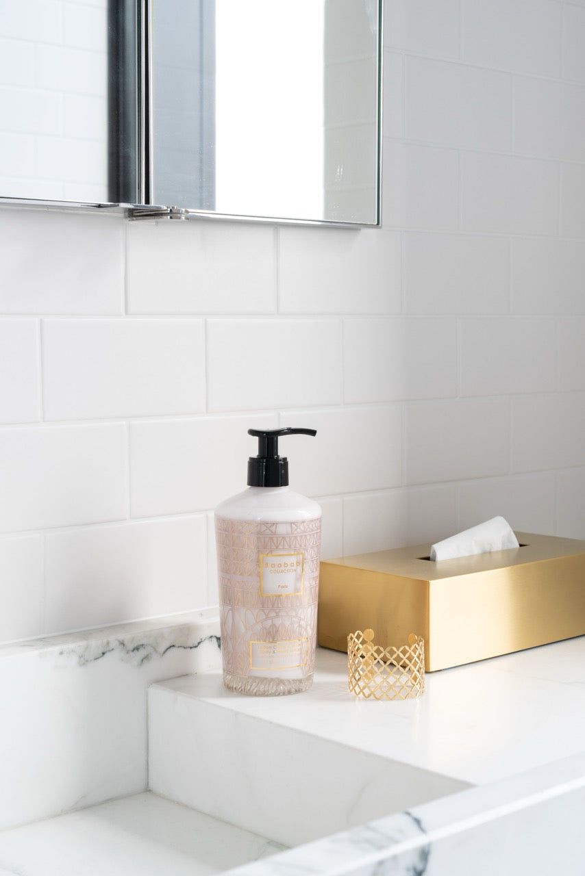 Front view of Paris Body & Hand Lotion bottle showcasing embossed pink glass and black pump on marble sink with gold tissue box