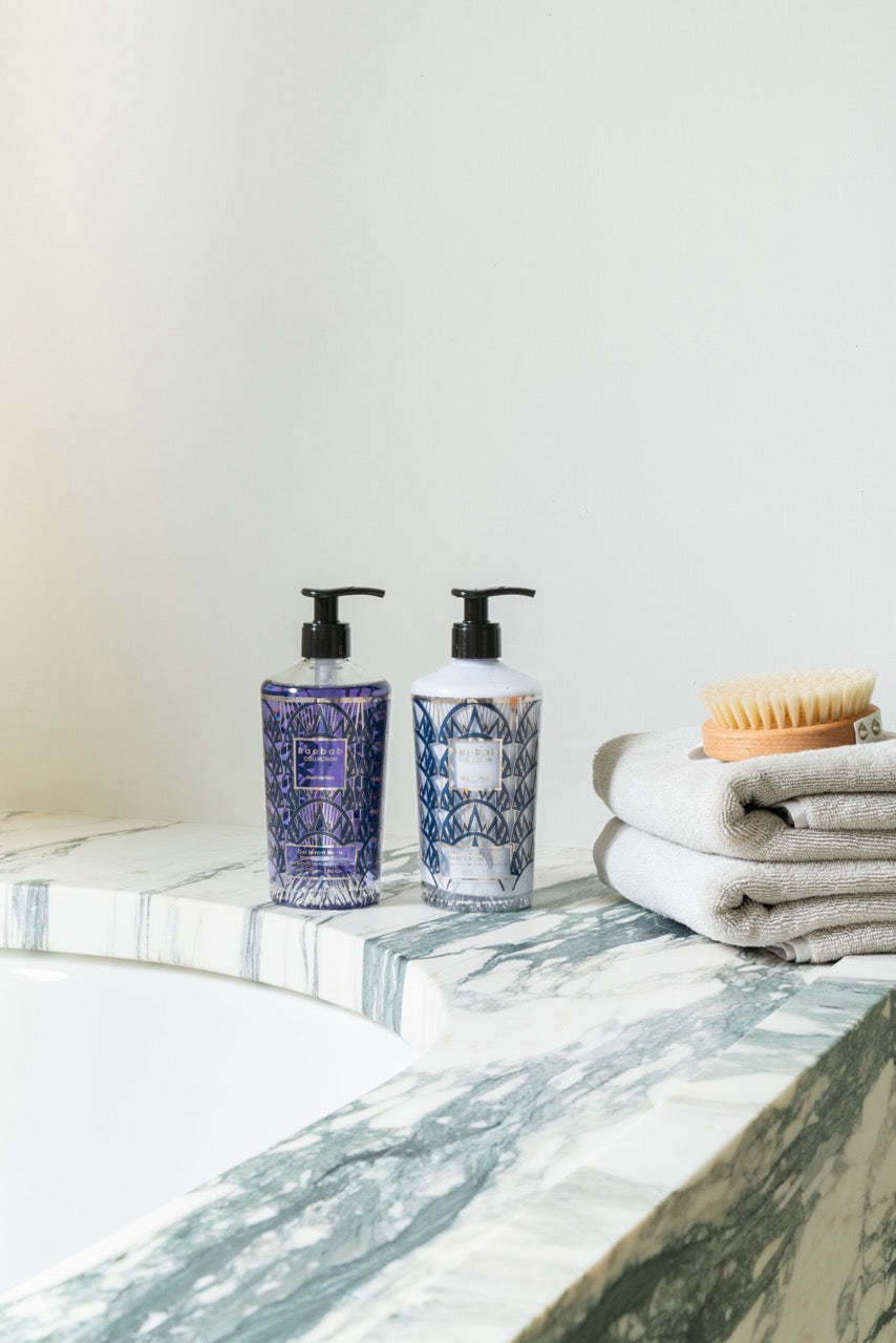 Two patterned pump bottles, hand wash and body lotion, on marble tub edge with towels and wooden brush