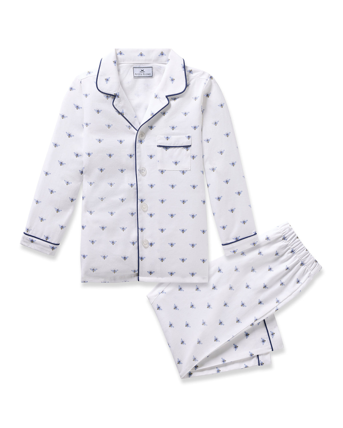 Petite Plumes Kids Twill Pajama in Buzzing Bees is a white cotton set with a long-sleeve button-up top and matching pants. Featuring navy blue trim on the collar, cuffs, and pocket, it is adorned with small blue buzzing bees for a charming contrast.