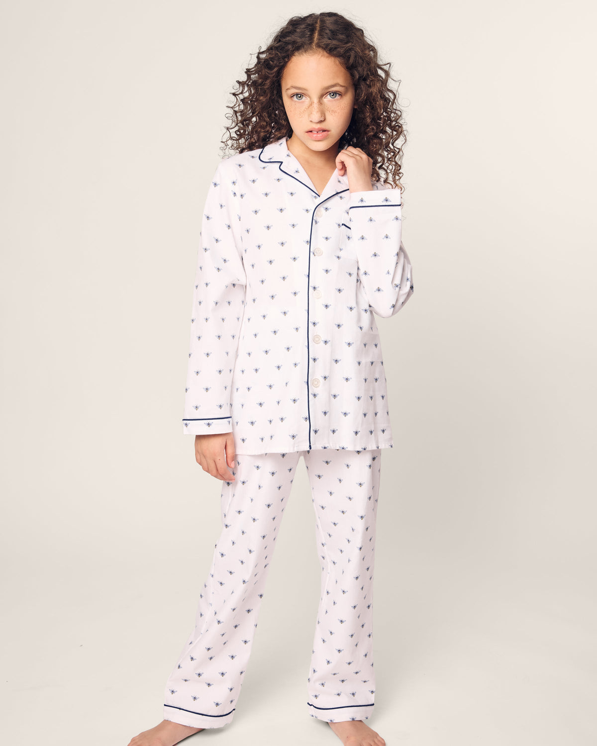 A young girl with curly hair models Petite Plumes Kids Twill Pajama in Buzzing Bees, featuring dark piping. She stands barefoot, hand by her face, against a light background, exuding comfort and charm.