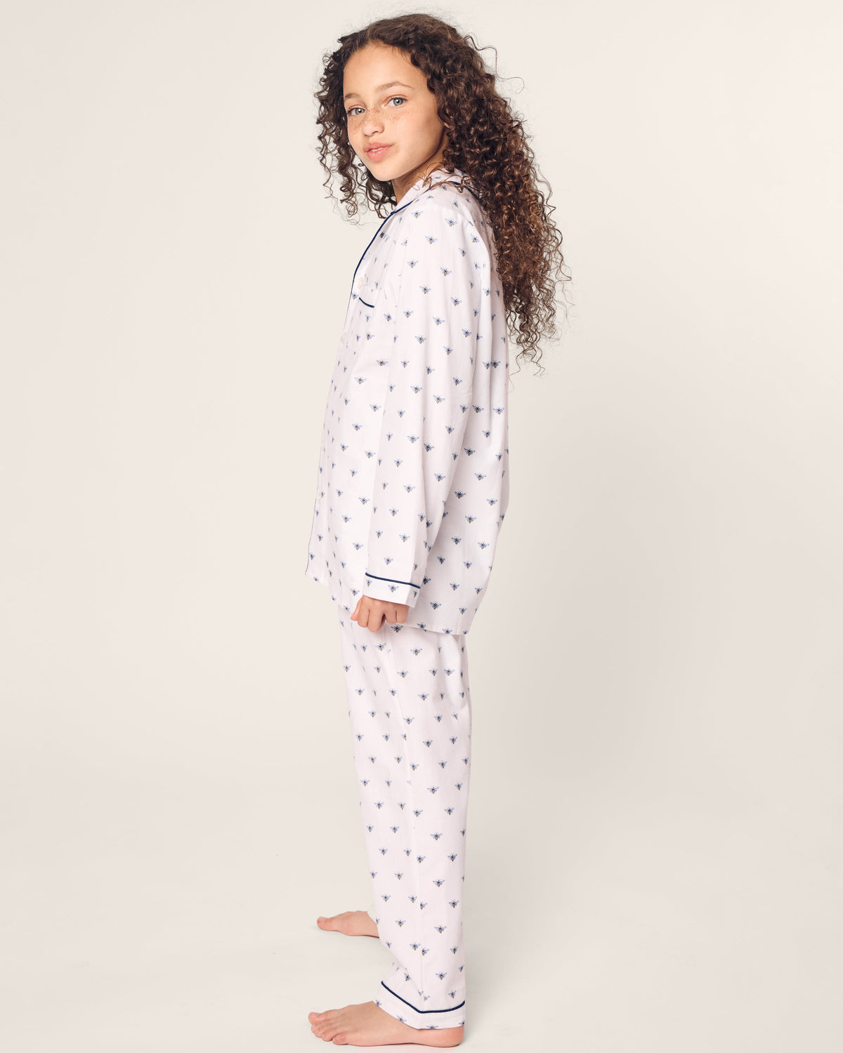 A young girl with curly hair stands barefoot, facing sideways, in Petite Plumes cozy Kids Twill Pajama set adorned with a subtle Buzzing Bees pattern and dark trim against a plain white background.