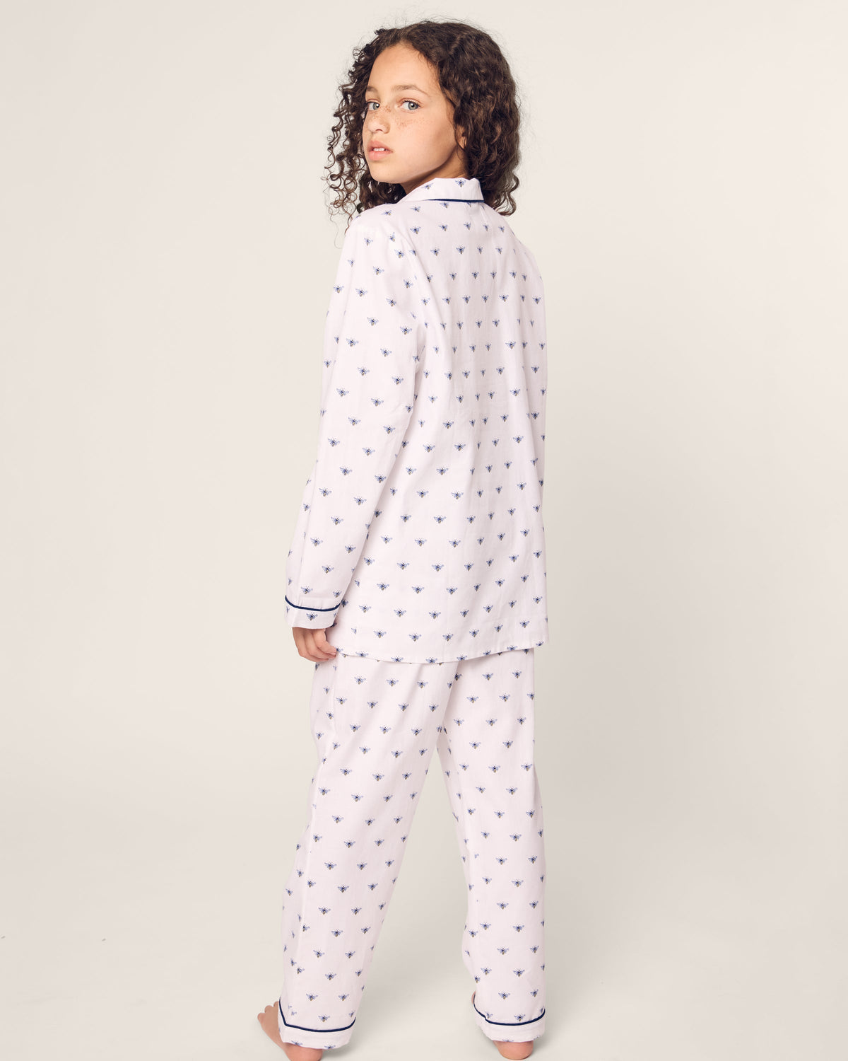 A curly-haired child stands barefoot in a comfortable Petite Plume Kids Twill Pajama set in Buzzing Bees, featuring a collar and dark trim on the sleeves and pants. The child gazes back at the camera against a plain background.