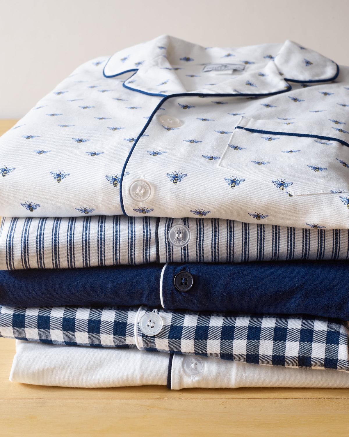 Four cotton pajama sets neatly rest on wood, starting with Petite Plumes Buzzing Bees print on white, followed by striped, solid navy, and checkered navy/white designs. Each set features unique button details and trim for charm.