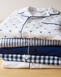 Four cotton pajama sets neatly rest on wood, starting with Petite Plumes Buzzing Bees print on white, followed by striped, solid navy, and checkered navy/white designs. Each set features unique button details and trim for charm.