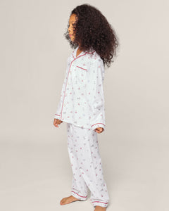 A child with curly hair stands sideways, wearing Petite Plumes Kids Twill Pajama in Coastal Charms. Made of yarn-dyed cotton, the pajamas feature small sailboat patterns and red piping on white fabric. The plain, light background highlights their charming sleepwear.